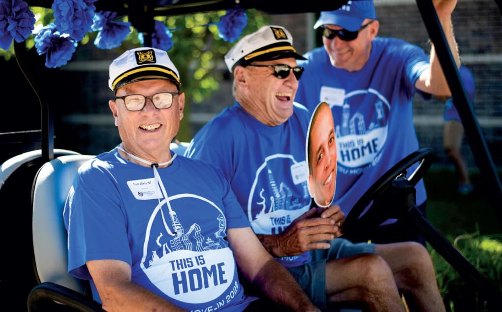 Two members of the Class of '72 sit in a golf cart wearing captains hats and blue t-shirts from move-in that read "This is home. Move-in 2022."